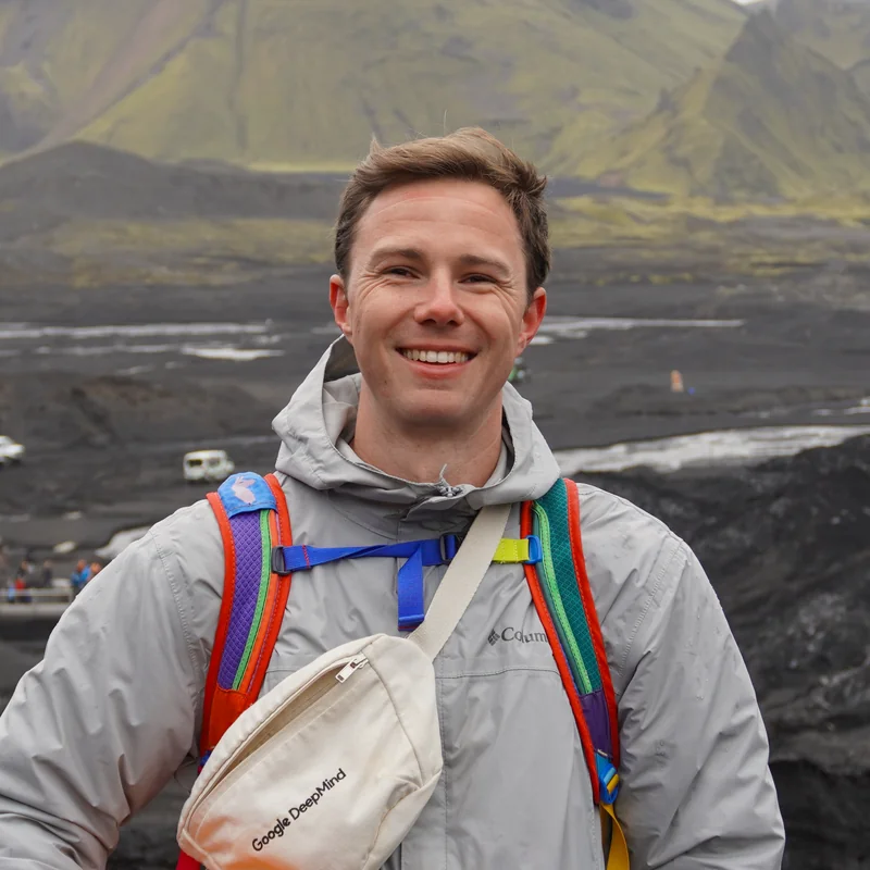 Sam Hopkins on a glacier hike in Iceland, smiling with an orange climbing helmet and a Google DeepMind bag